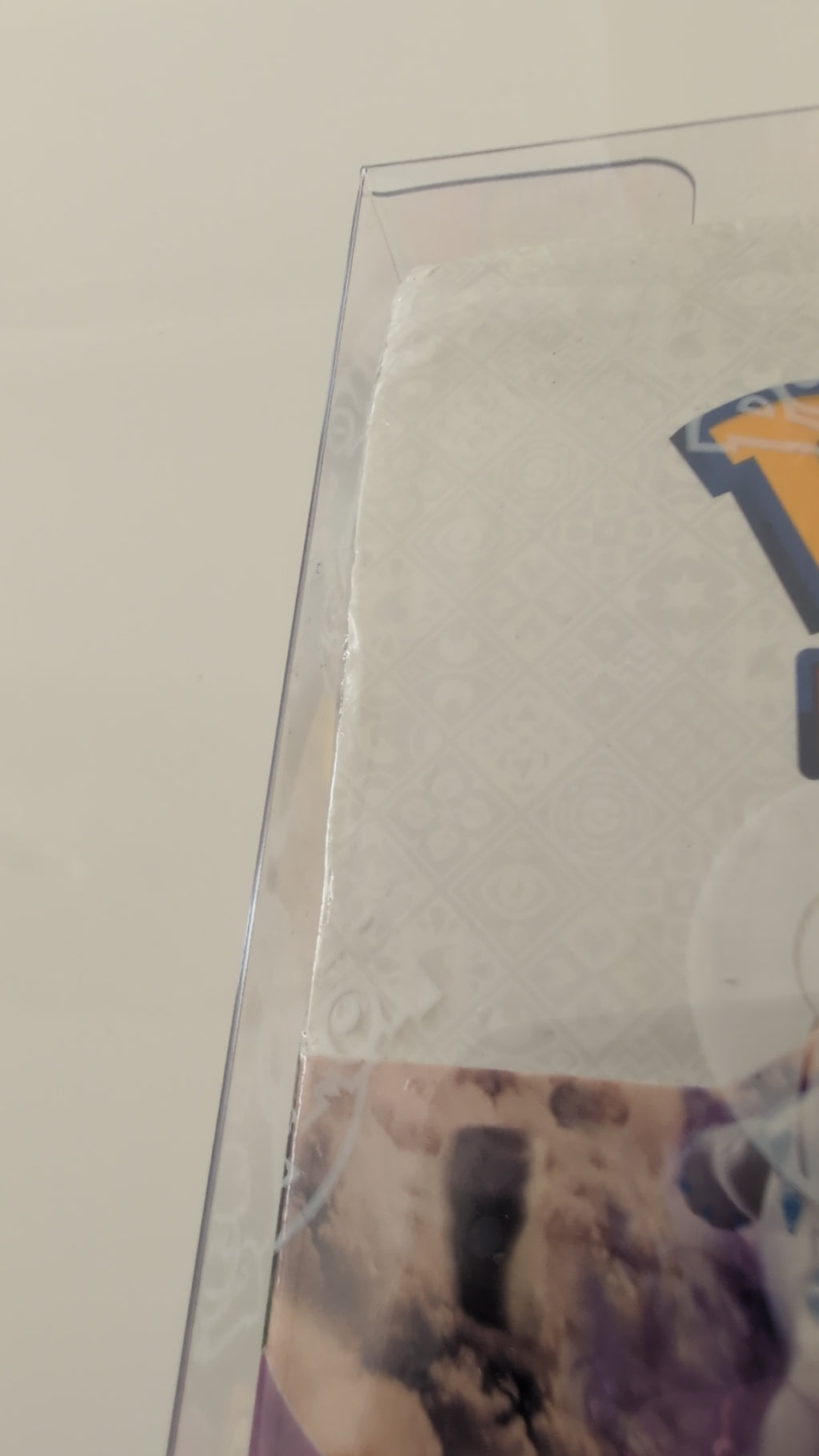 A close-up of a clear plastic protective sleeve or case over a printed card or insert, showing part of a white patterned section and a colorful graphic with yellow, blue, and purple tones.